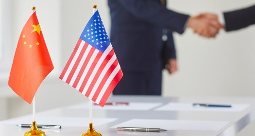A conference table with the flags of the US and China, with two men shaking hands in the background.