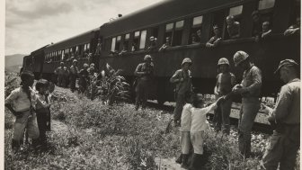 A black and white photo of U.S. Soldiers standing next to a stopped train, handing out candy to South Korean children.