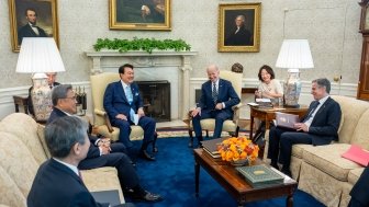 A group of diplomats surrounding Presidents Yoon and Biden in the Oval Office, the group is laughing.