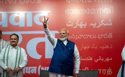 Narendra Modi stands in front of a red background, holding up his hand in a victory sign.